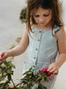 A young girl with long brown hair looks down while touching pink flowers near a plant. She is wearing a sleeveless, light blue and white vertically striped button-front dress.