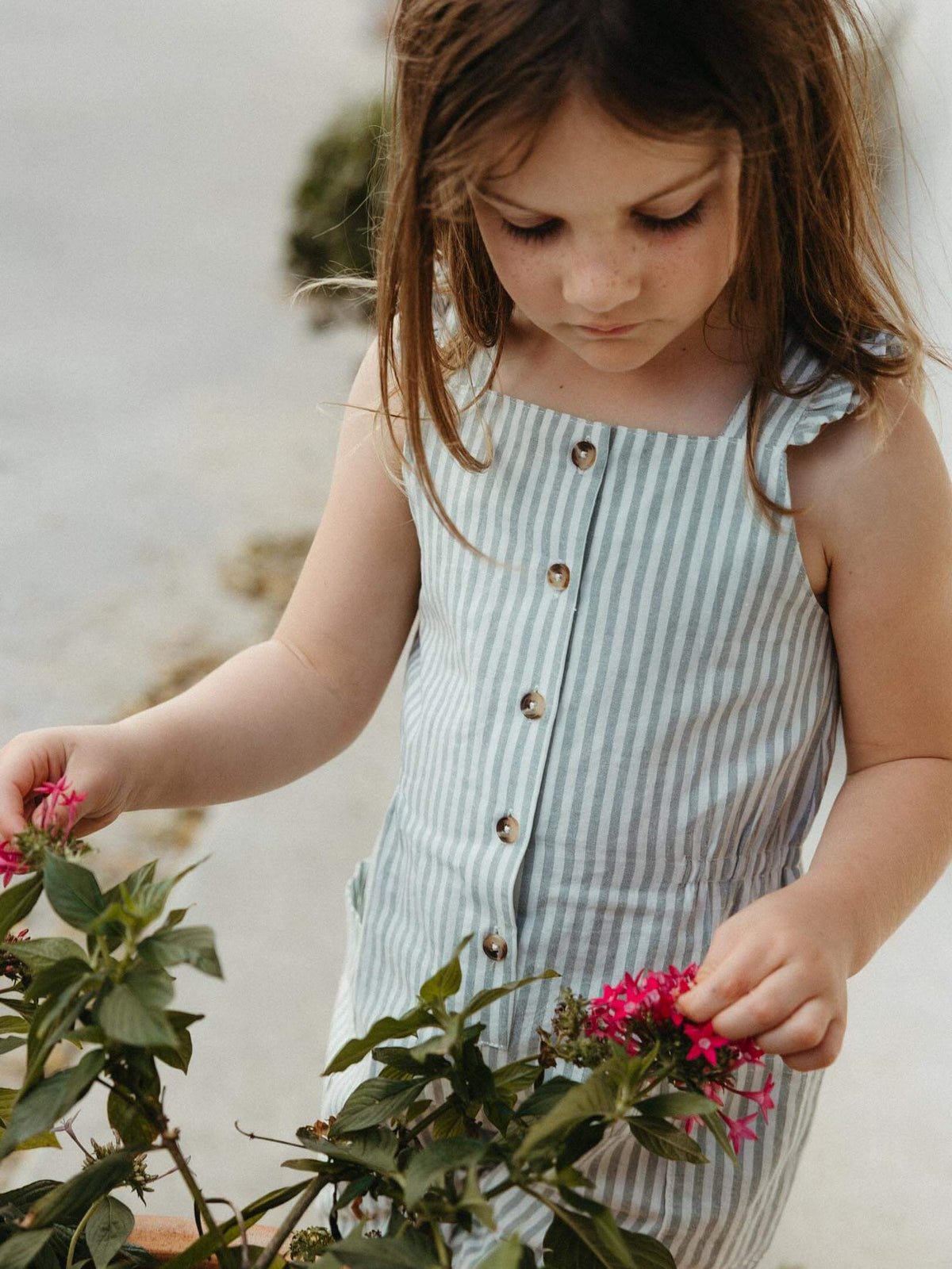 A young girl with long brown hair looks down while touching pink flowers near a plant. She is wearing a sleeveless, light blue and white vertically striped button-front dress.