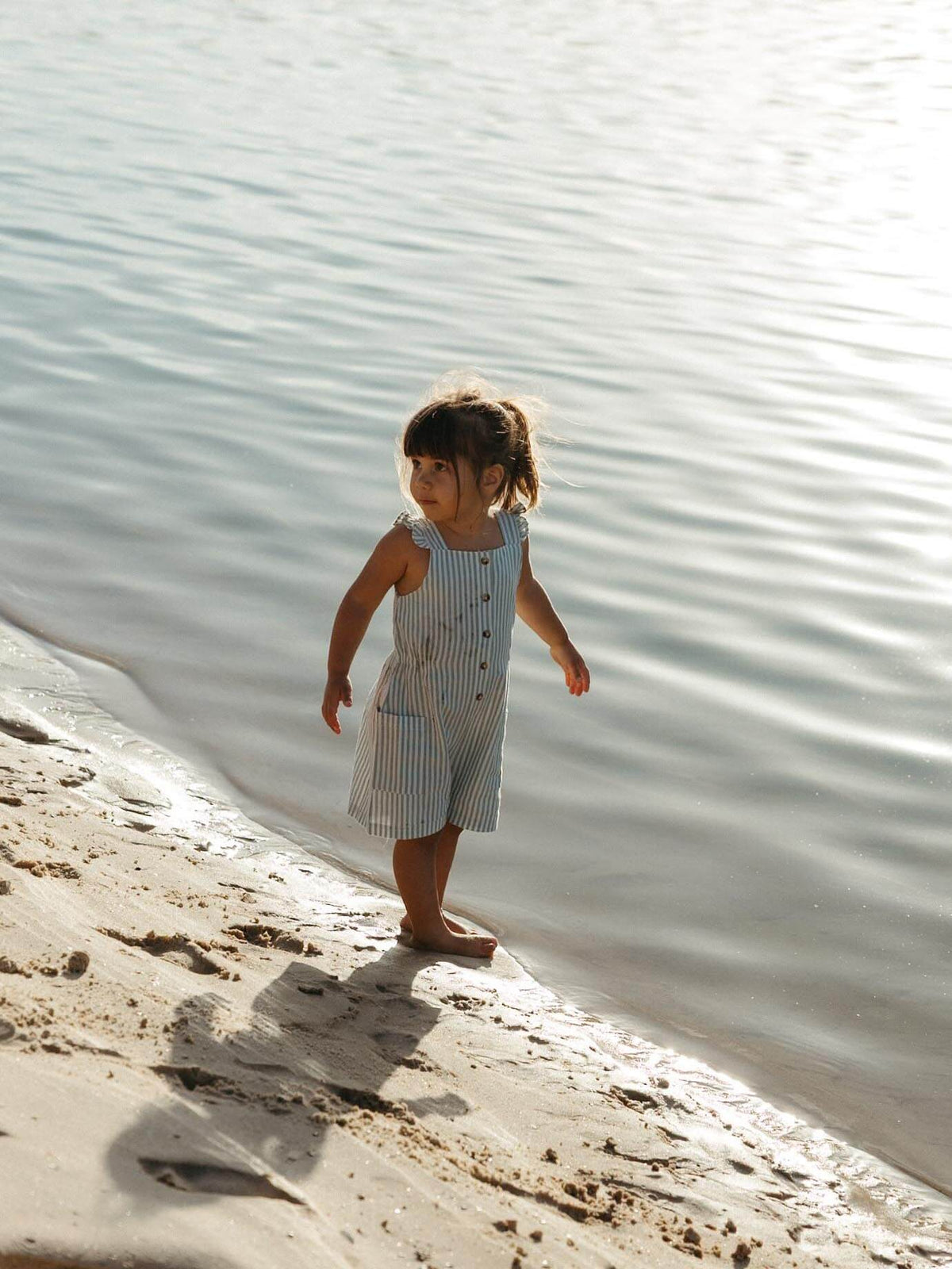 A young child wearing a blue and white striped sleeveless jumpsuit stands barefoot at the edge of a calm body of water on a sandy shore.