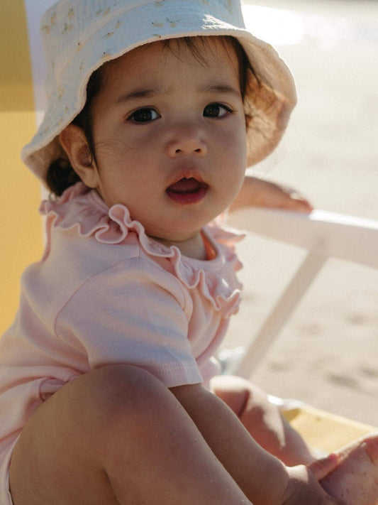 A baby wearing a light pink ruffled short-sleeve top and a white sun hat with small gold stars, sitting outdoors on a beach chair.