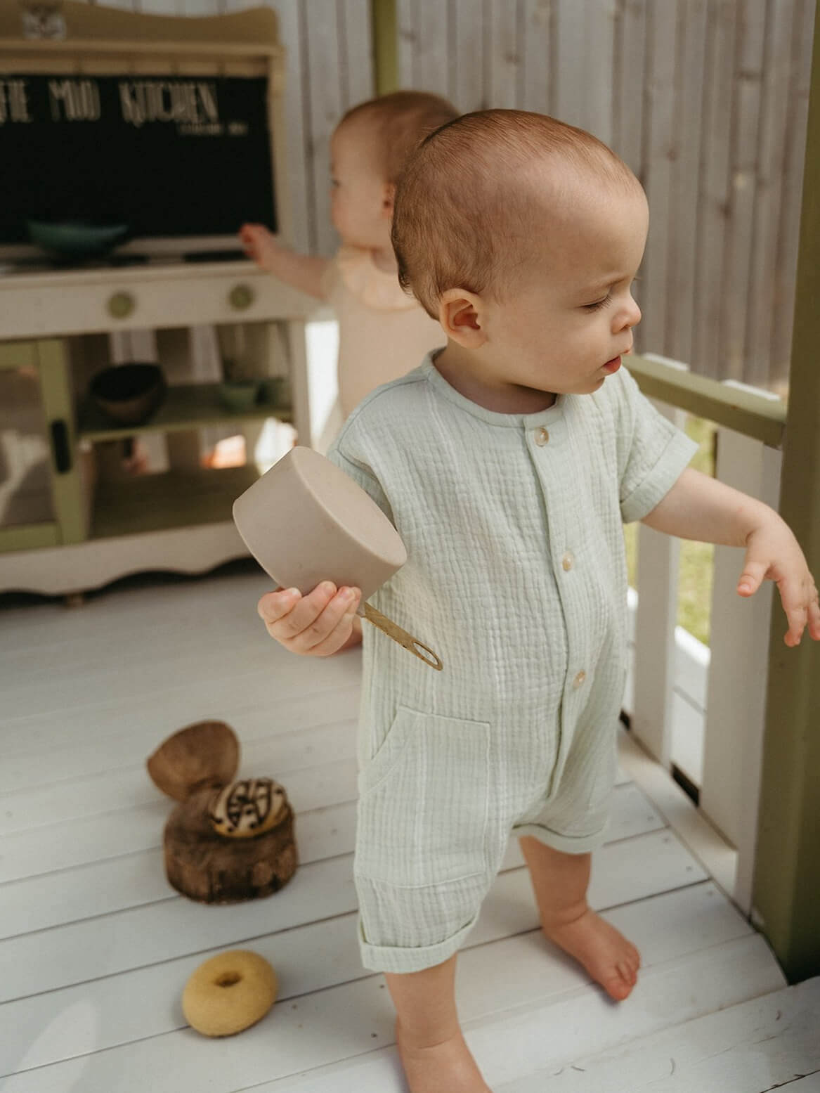 Child in a light green outfit holding a toy kitchen utensil on a wooden floor.