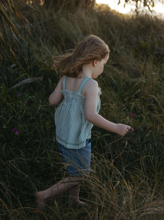 A young child walking through tall grass outdoors at sunset, wearing a light blue and white striped sleeveless top and denim shorts.