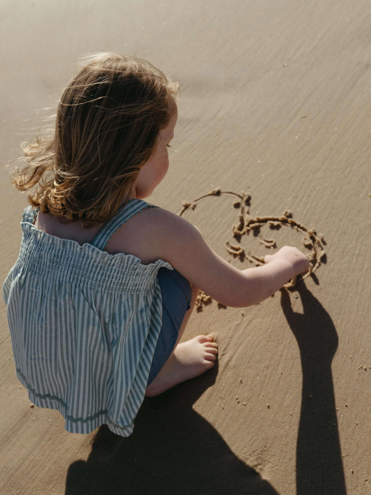 A young child wearing a blue and white striped romper crouches on a sandy beach, drawing a heart in the sand with their hand.