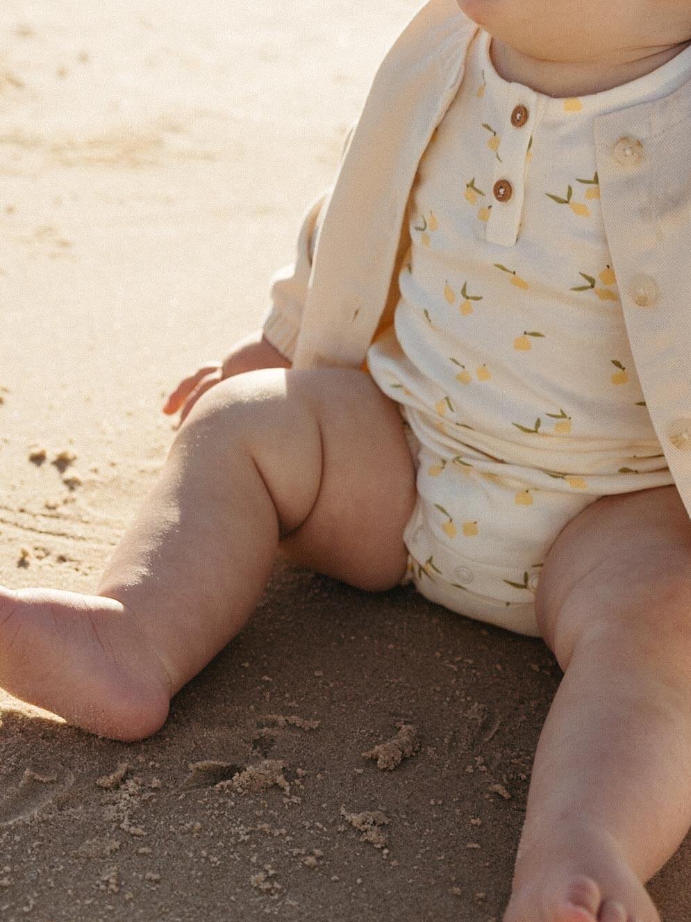 A close-up of a baby sitting on sand, wearing a cream-colored romper with a small yellow floral print and a matching cream cardigan.