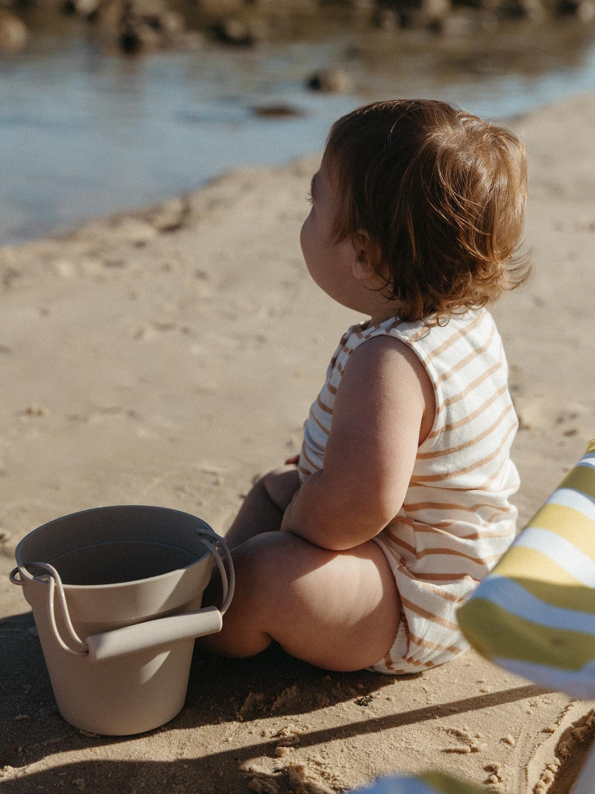 A toddler sits on a sandy beach next to a small bucket, wearing a sleeveless cream and tan striped romper. The background shows water and rocks in soft focus.