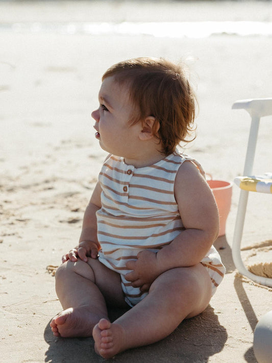 A baby sitting on a sandy beach wearing a sleeveless cream and tan striped romper, looking off-camera. A small white beach chair is partially visible on the right.