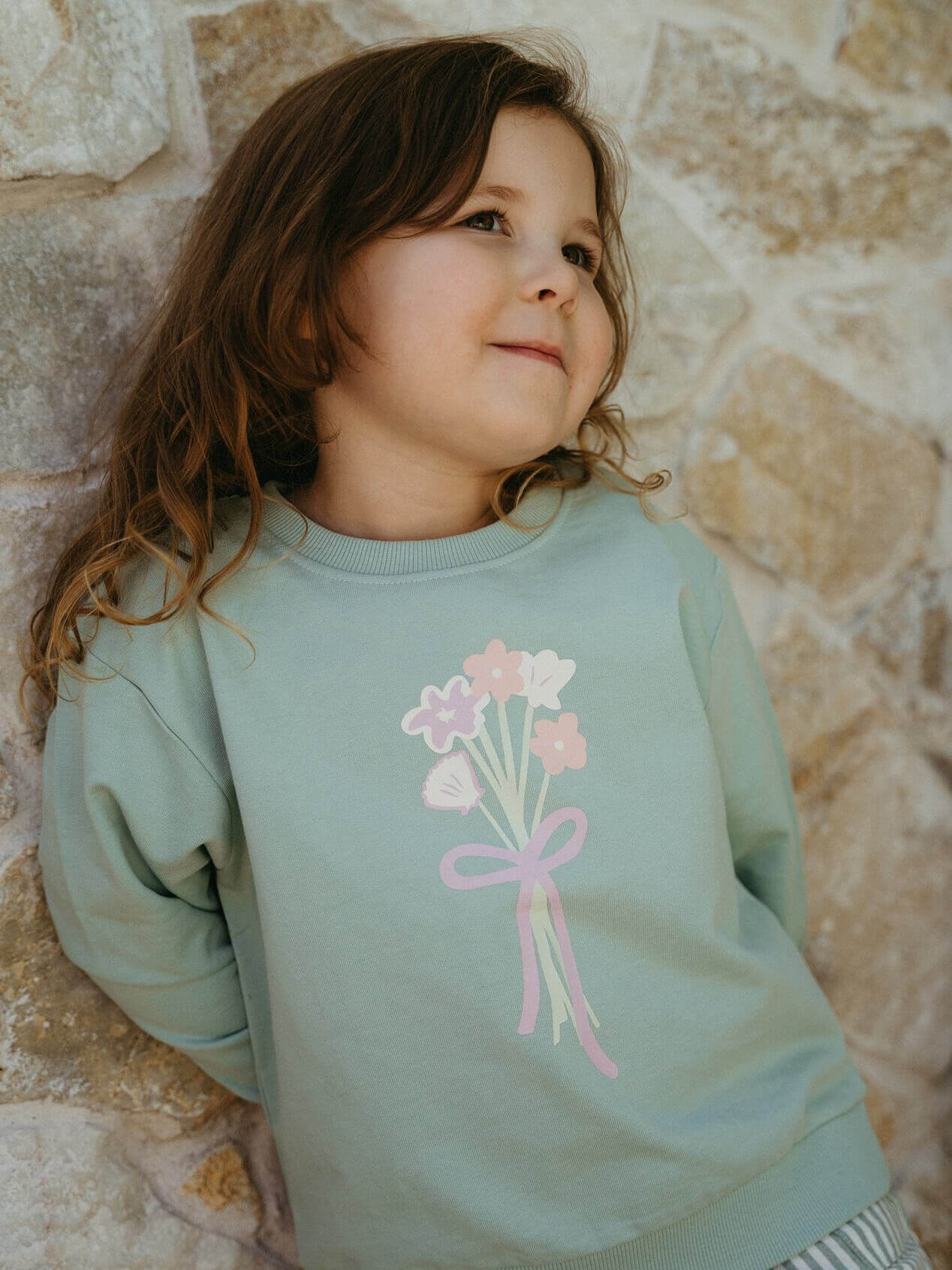 Child wearing a light blue sweatshirt with a floral design against a stone wall.