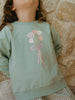 Light green sweatshirt with floral design worn by a child against a stone wall background