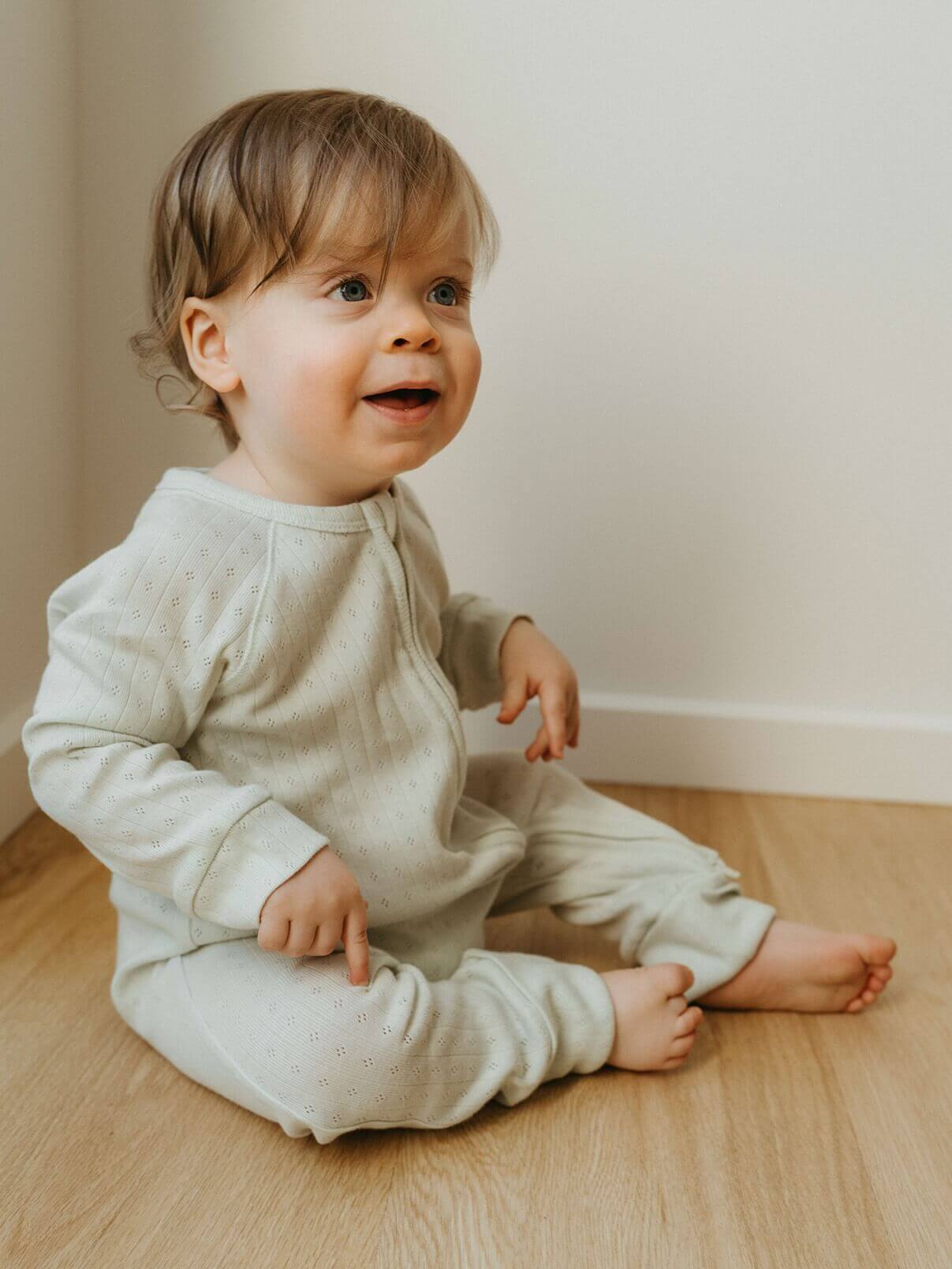 Child wearing a light gray onesie sitting on a wooden floor.