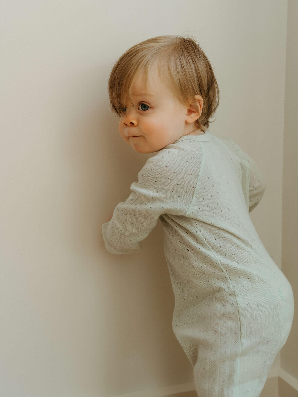 Baby in a white outfit standing against a plain wall