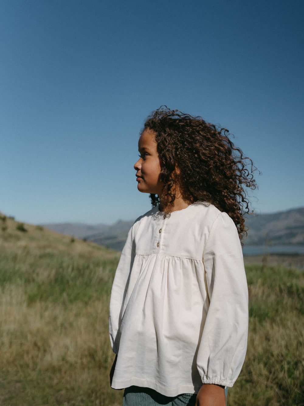 Young girl in a white blouse standing in a grassy field with mountains in the background Three Button Blouse - Ecru Girls