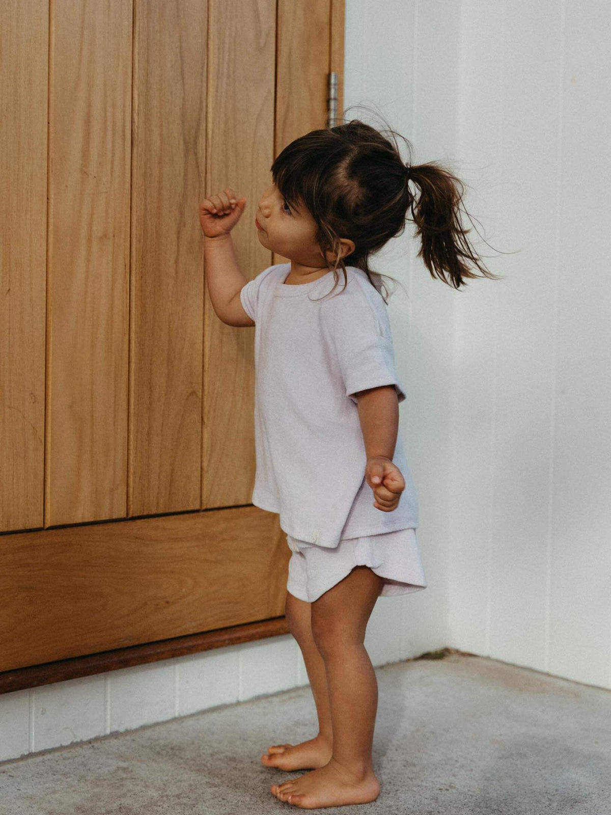 A toddler girl with a ponytail stands barefoot in a corner, wearing a light lavender short-sleeve t-shirt and matching shorts. She is positioned next to a light wood paneled door and a white wall.
