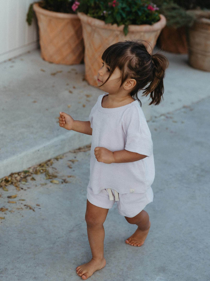 A toddler girl walking barefoot on a concrete patio wearing a light gray short-sleeve romper with matching shorts.