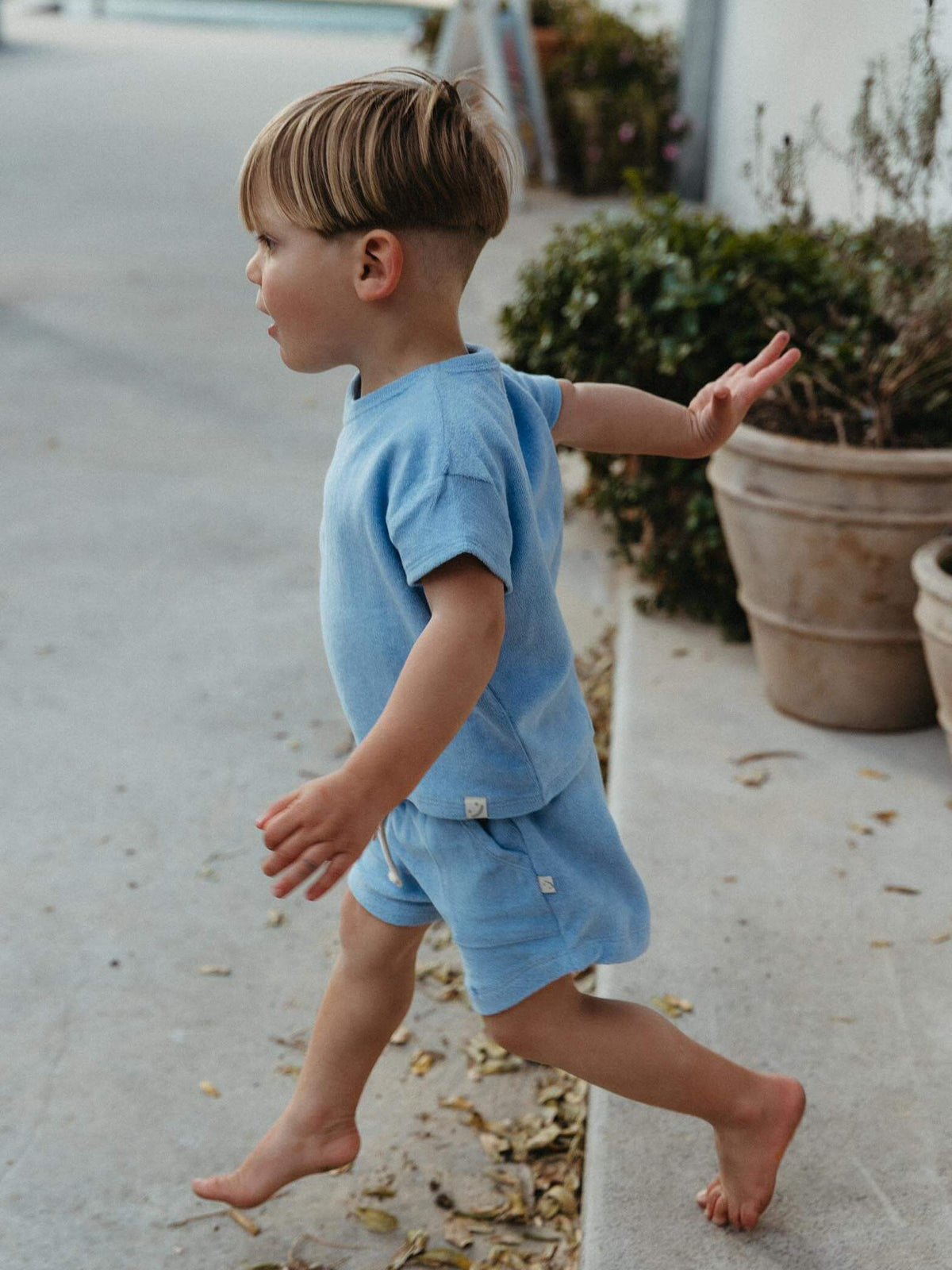 A young child wearing a light blue short-sleeve t-shirt and matching shorts walks barefoot along a concrete path outdoors.