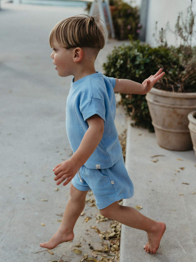 A young child wearing a light blue short-sleeve t-shirt and matching shorts walks barefoot along a concrete path outdoors.