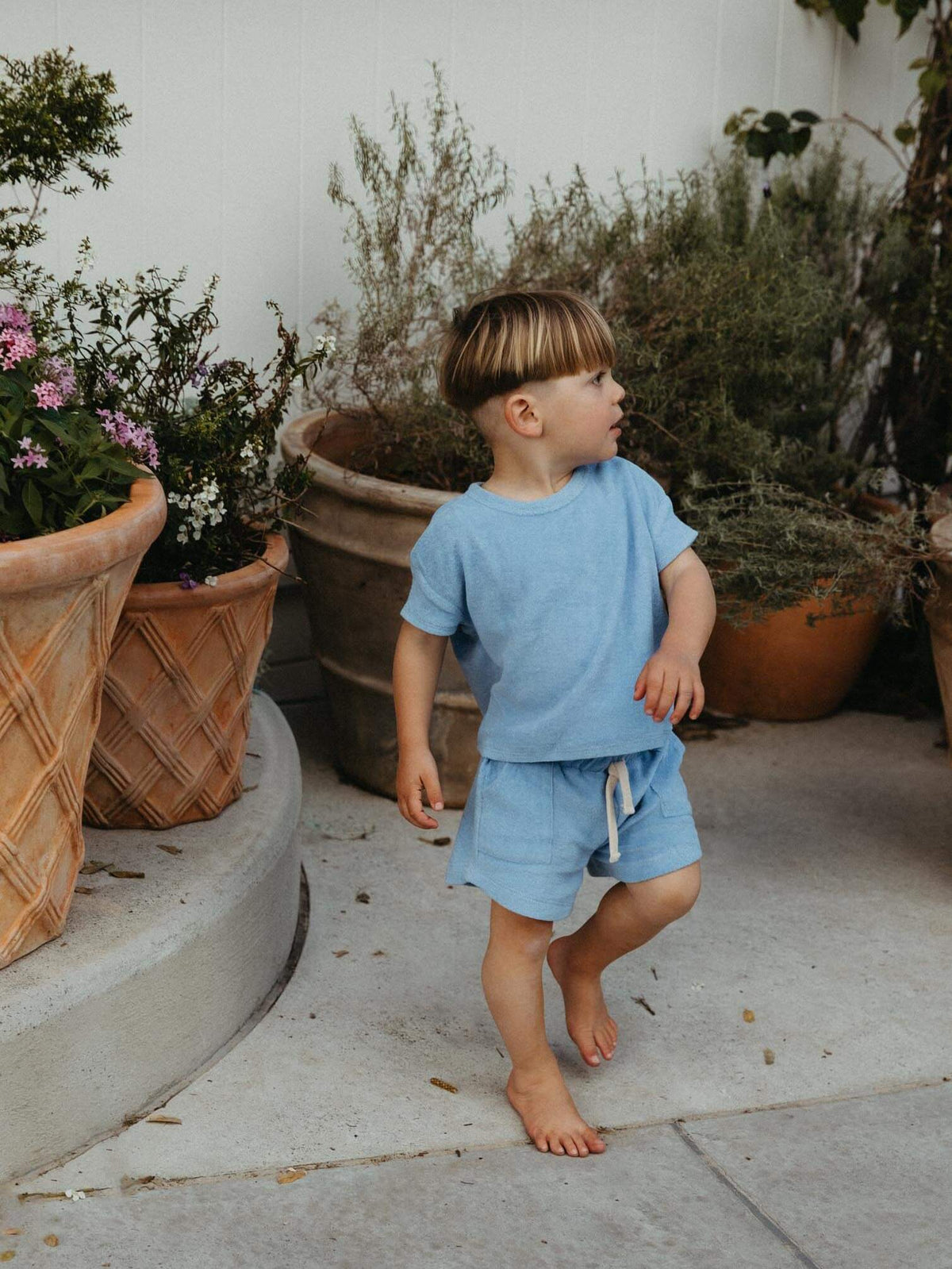 A young child wearing a light blue short-sleeve t-shirt and matching shorts walks barefoot on a concrete patio next to large potted plants.