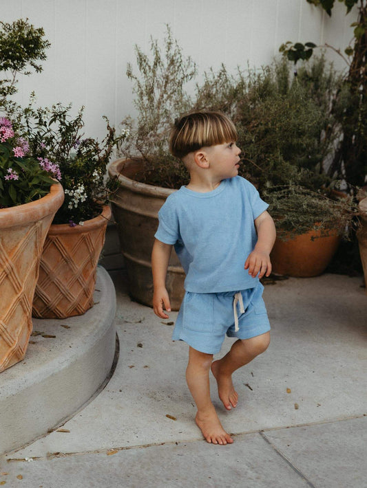 A young child wearing a light blue short-sleeve t-shirt and matching shorts walks barefoot on a concrete patio next to large potted plants.