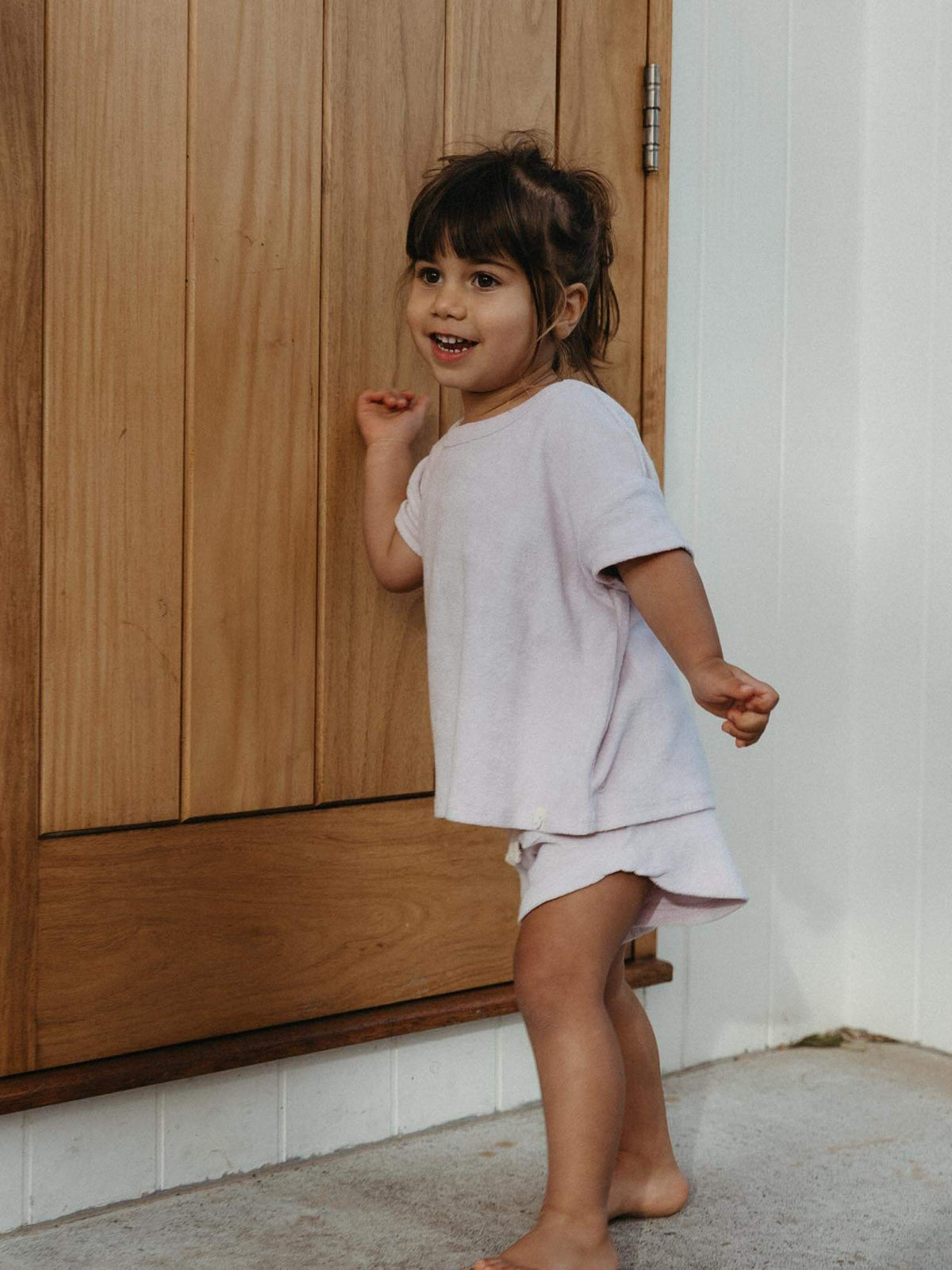 A young child wearing a light pink t-shirt and matching shorts stands barefoot in front of a wooden door and white wall.