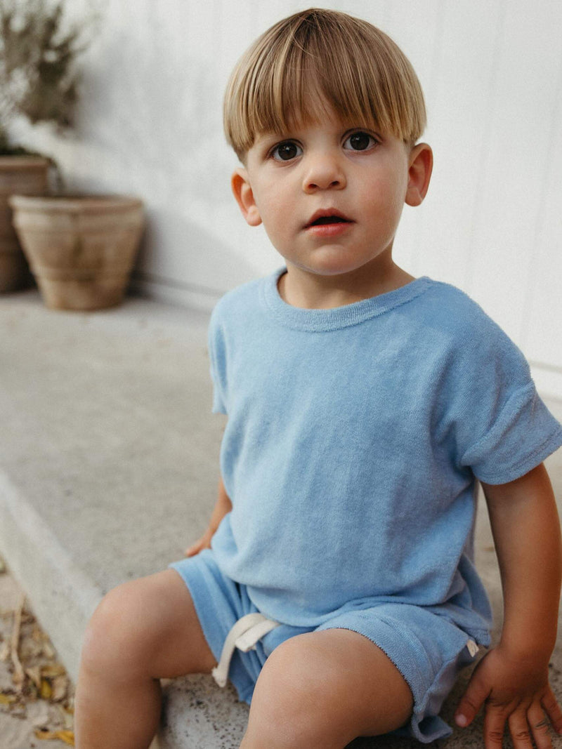 A young child wearing a light blue short-sleeve t-shirt and matching blue shorts with a white drawstring, sitting on a concrete ledge in an outdoor setting.