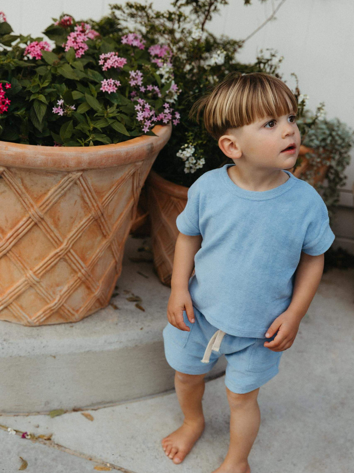 A toddler standing barefoot outdoors next to large potted flowers, wearing a light blue short-sleeve t-shirt and matching light blue shorts with a white drawstring.