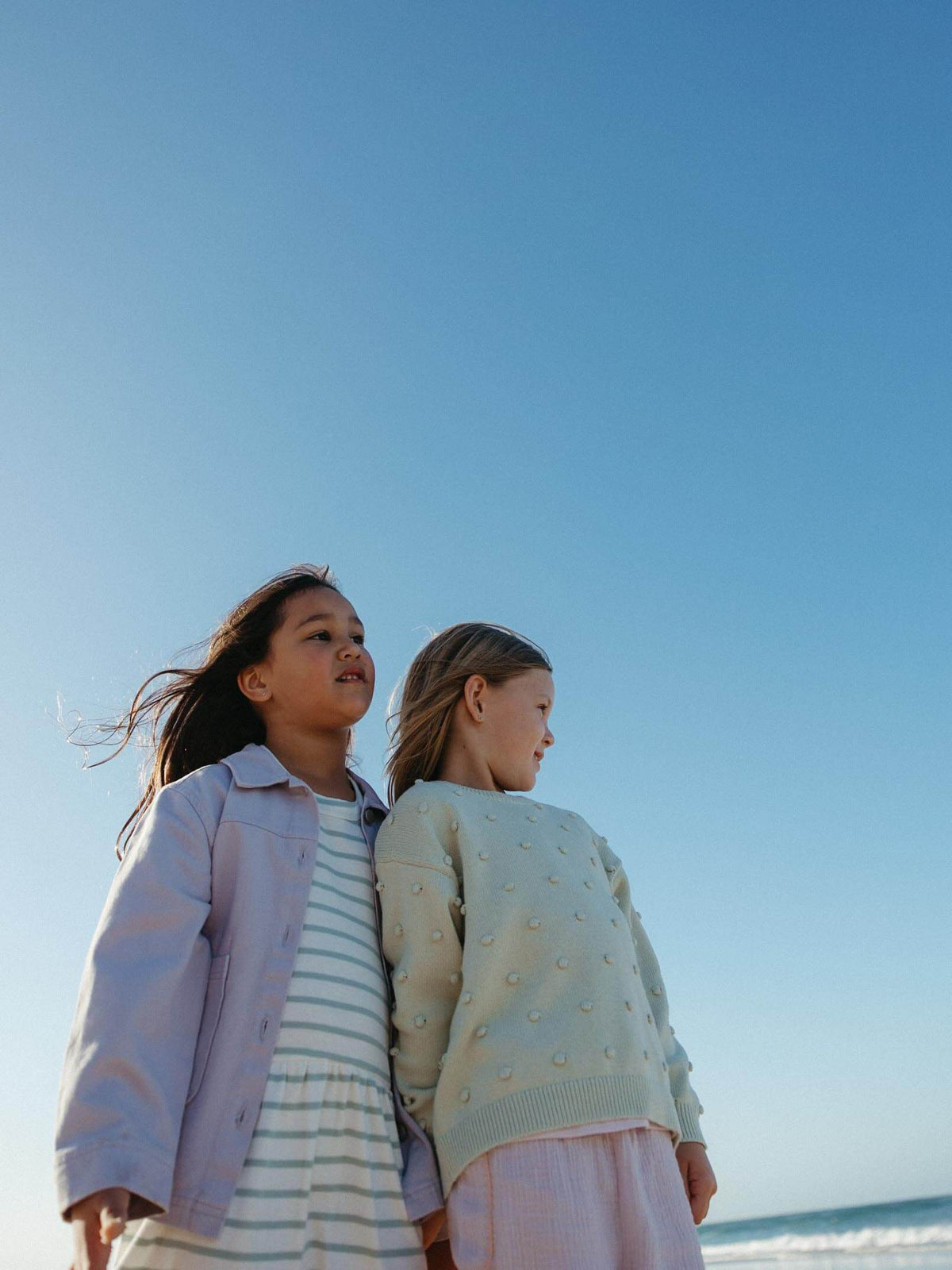 Two young girls standing side-by-side on a beach under a clear blue sky. The girl on the left wears a light purple button-up shirt over a white and blue striped dress, and the girl on the right wears a pale green textured knit sweater with a pink skirt.
