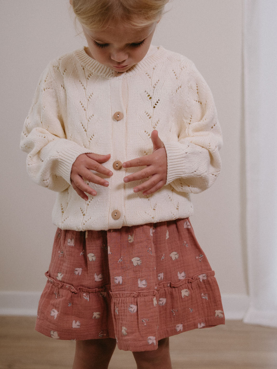 Young child wearing a cream cardigan and patterned pink skirt buttoning the sweater Woven Skirt - Bird And Blossom Rosehip Girls