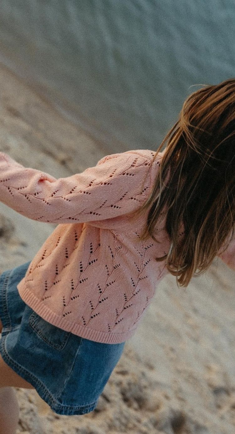 A child wearing a light pink knit sweater with a textured leaf pattern and blue denim shorts, photographed outdoors against a sandy, blurred background.