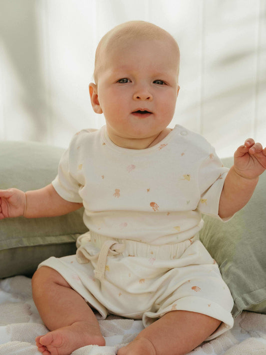 Baby in a white outfit with small patterns sitting on a light-colored surface.